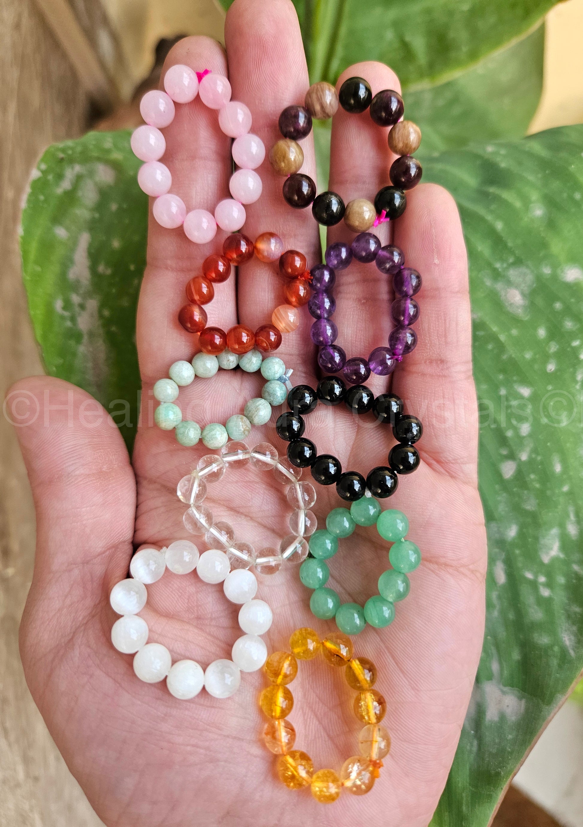 Hand holding multiple beaded bracelets of various colors against a green leafy background