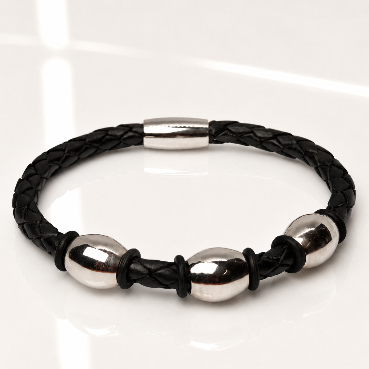 A black braided leather bracelet with three polished silver steel oval beads and a matching silver clasp, displayed on a clean white reflective surface.