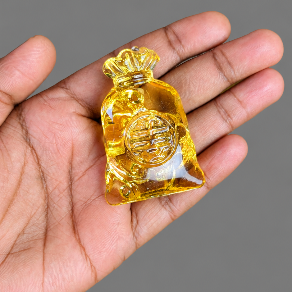 A translucent yellow glass money bag pendant held in a person's hand against a solid gray background.