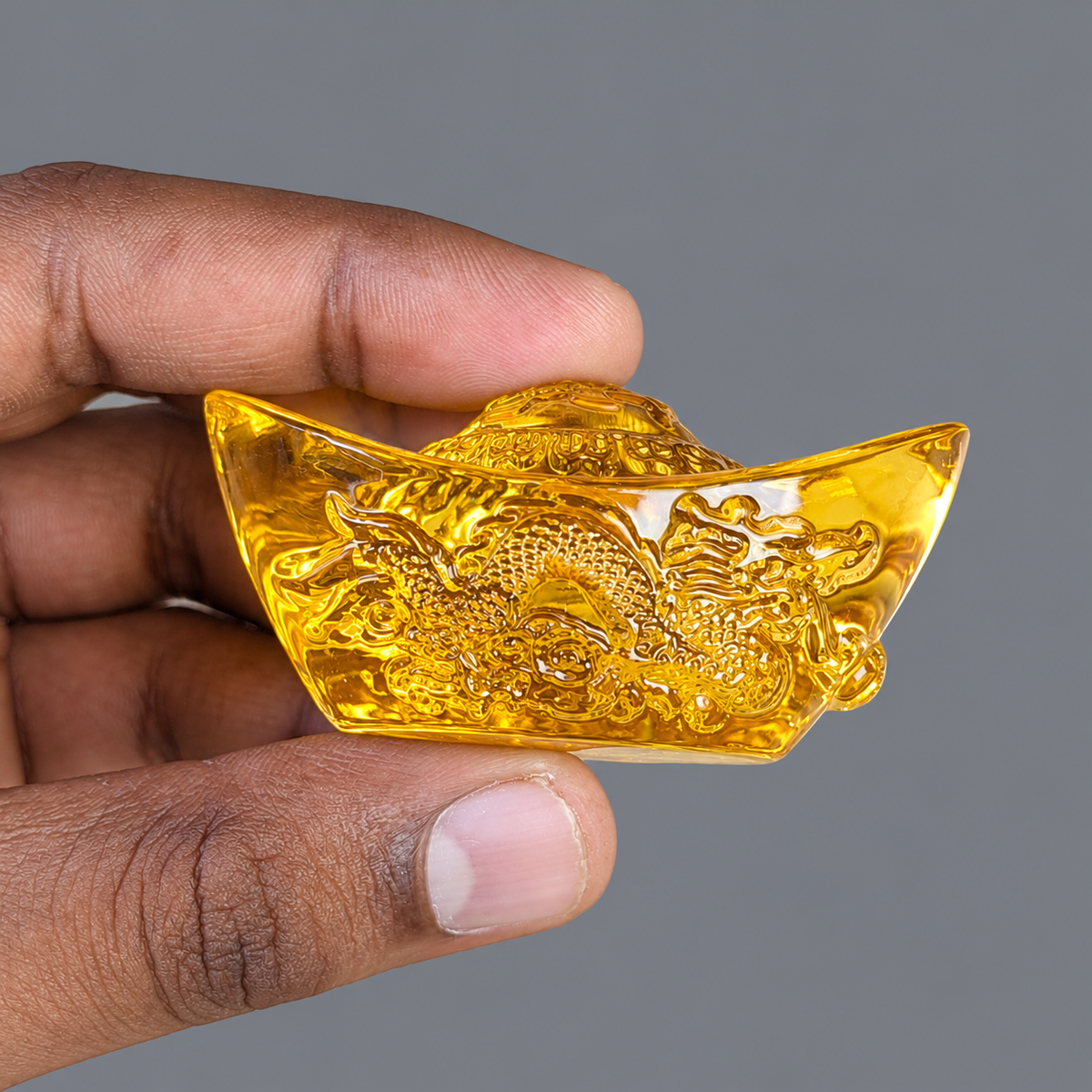 A hand holding a small translucent golden ingot-shaped ornament with an embossed dragon design, shown against a plain gray background.