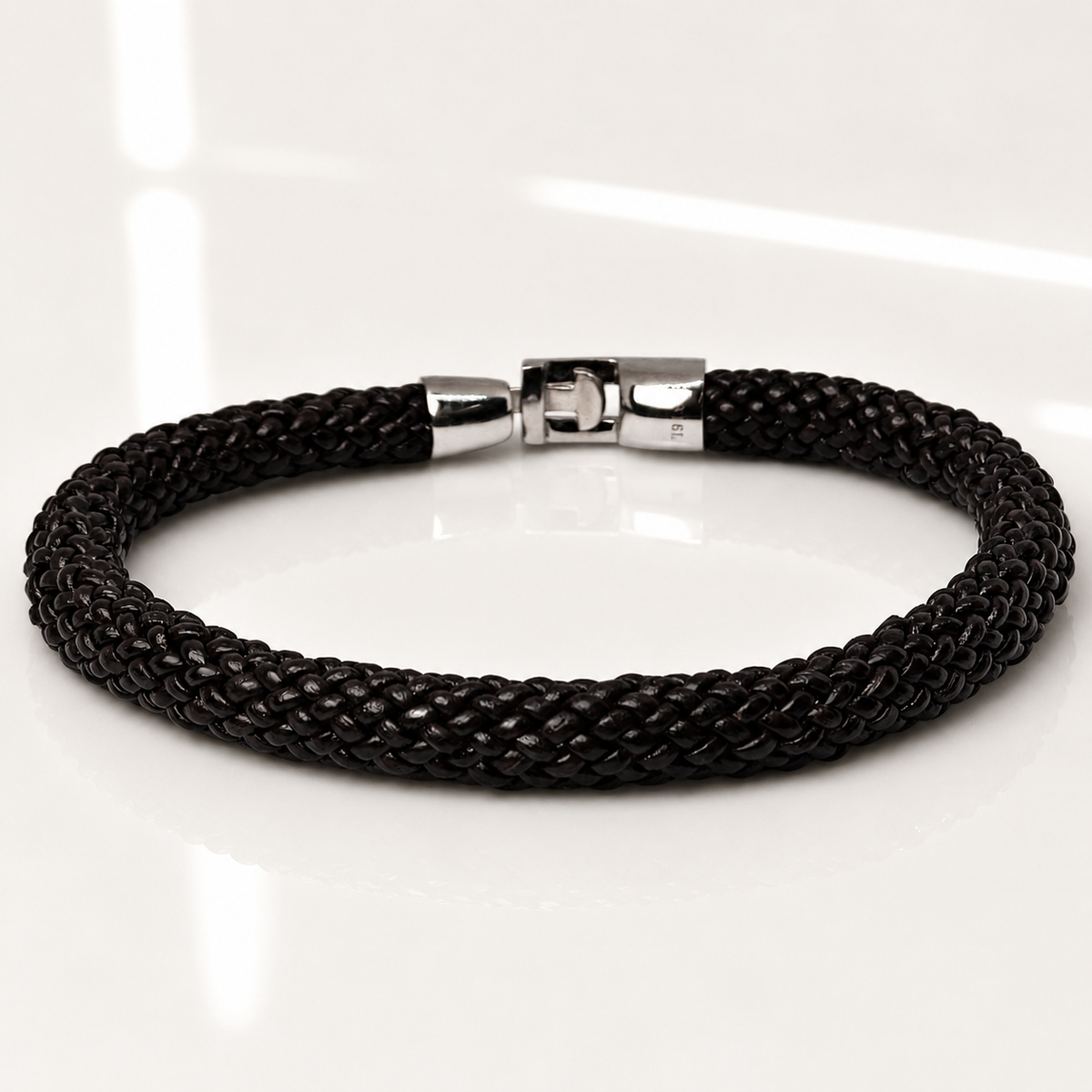 A black braided cord bracelet with a polished silver-toned clasp, displayed on a clean white reflective surface.