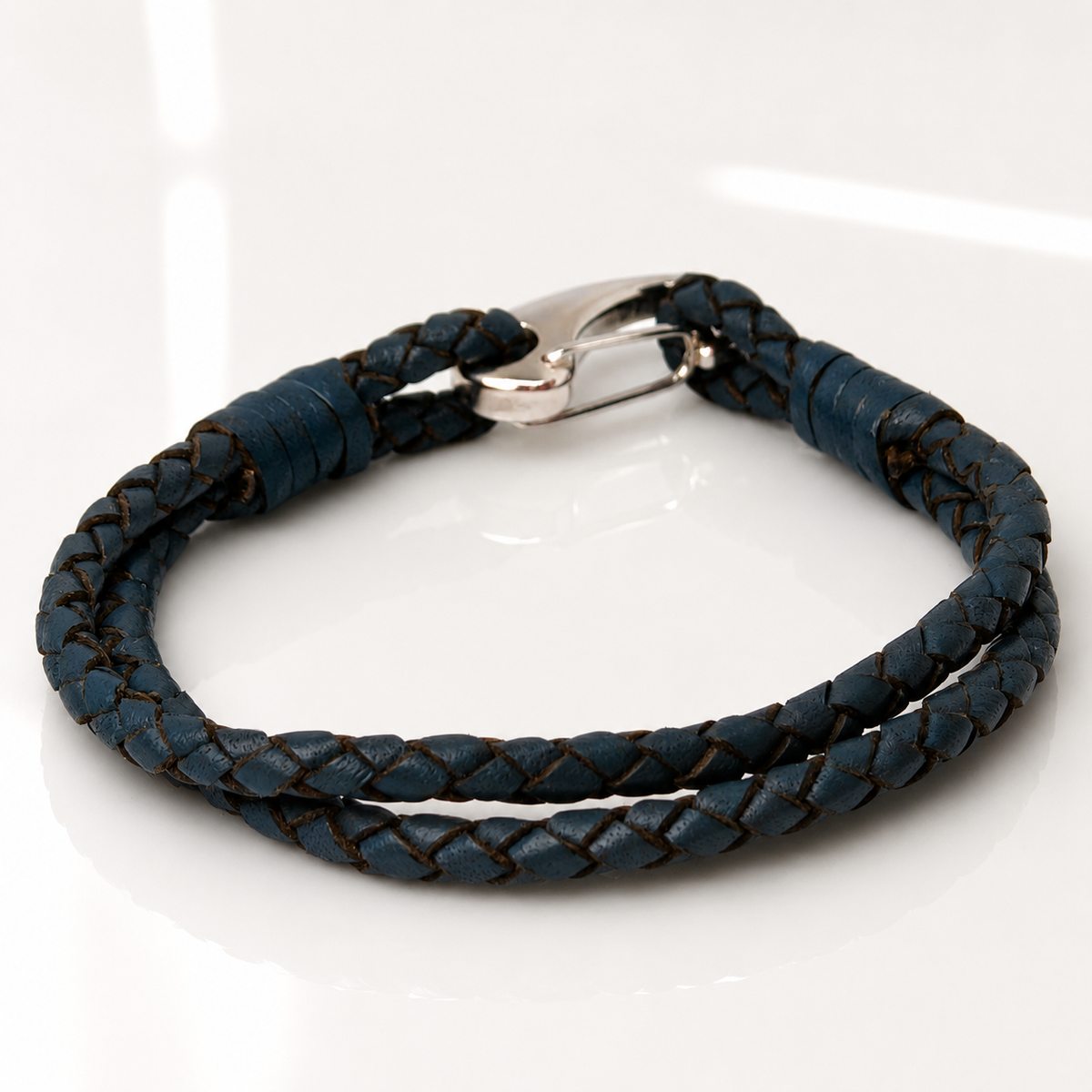 A dark blue braided leather bracelet with a silver metal clasp, displayed on a clean white reflective surface.