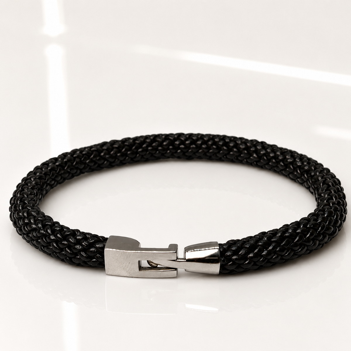 A black braided cord bracelet with a polished silver-tone metal clasp, displayed on a clean white reflective surface.