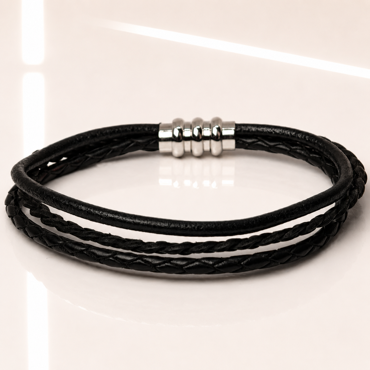 A black multi-strand braided cord bracelet with a polished silver-toned bead clasp, displayed on a clean white reflective surface.