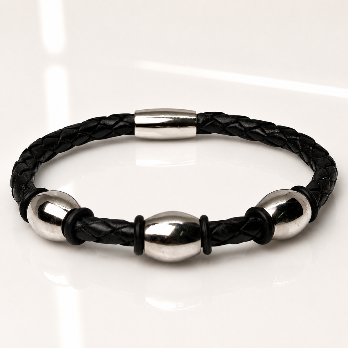 A black braided leather bracelet with polished silver steel beads and a silver clasp, displayed on a clean white reflective surface.