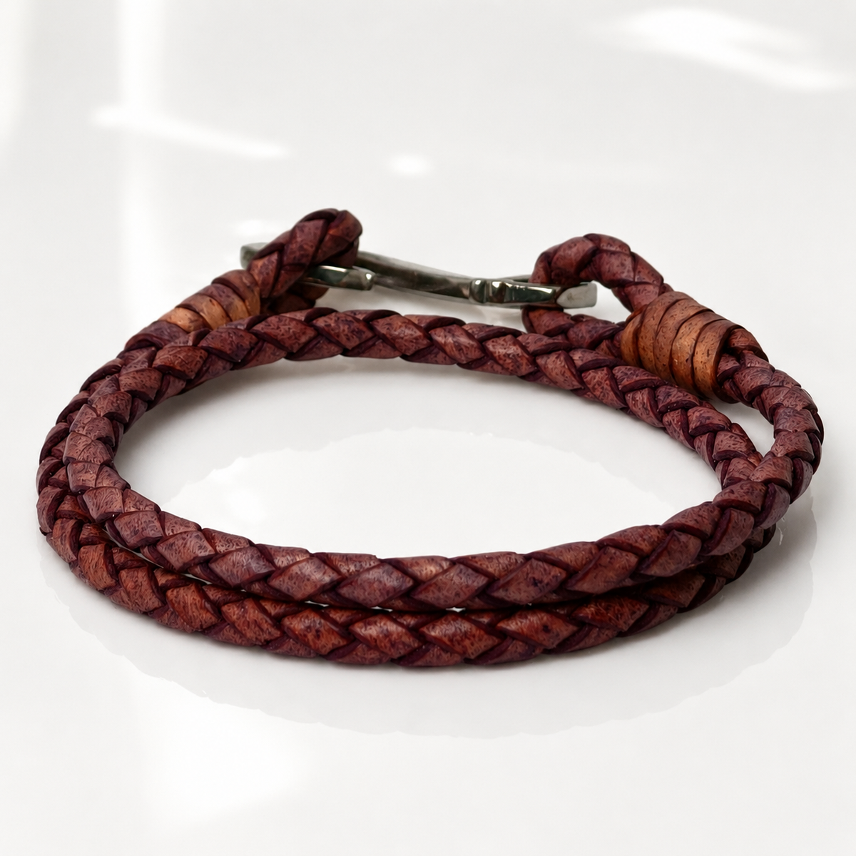 A braided leather bracelet in a reddish-purple hue with a silver metal clasp, displayed on a clean white reflective surface.
