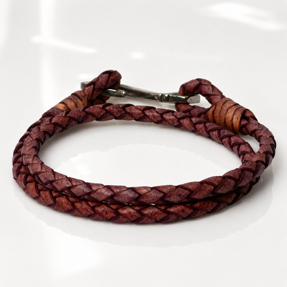 A braided leather bracelet in a reddish-purple hue with a silver metal clasp, displayed on a clean white reflective surface.