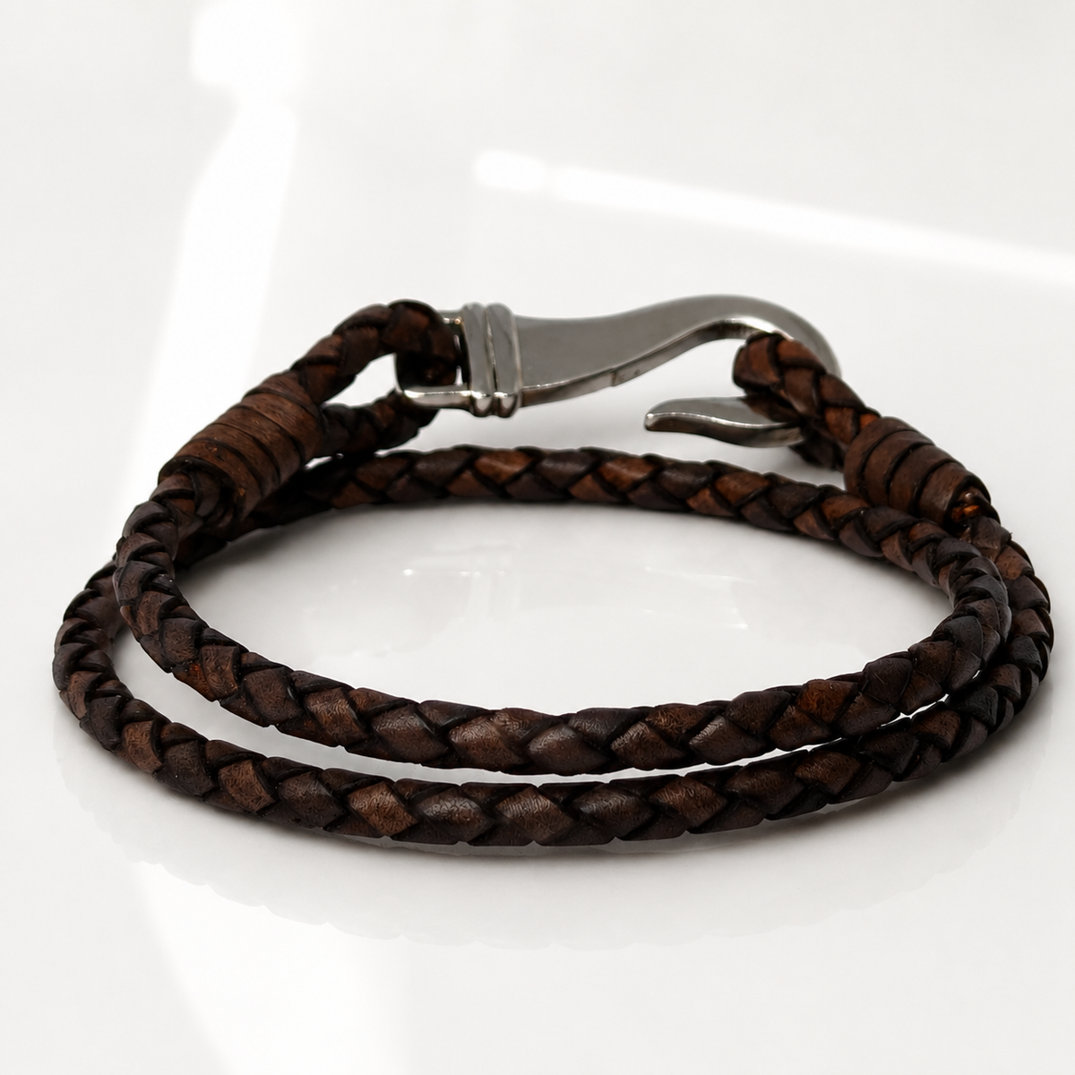 A dark brown braided leather bracelet with a silver-toned metal lobster clasp, displayed on a clean white reflective surface.
