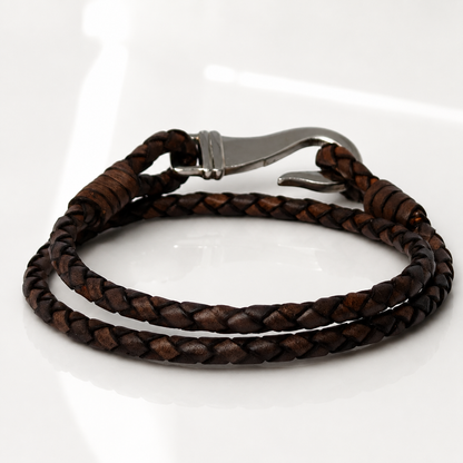 A dark brown braided leather bracelet with a silver-toned metal lobster clasp, displayed on a clean white reflective surface.