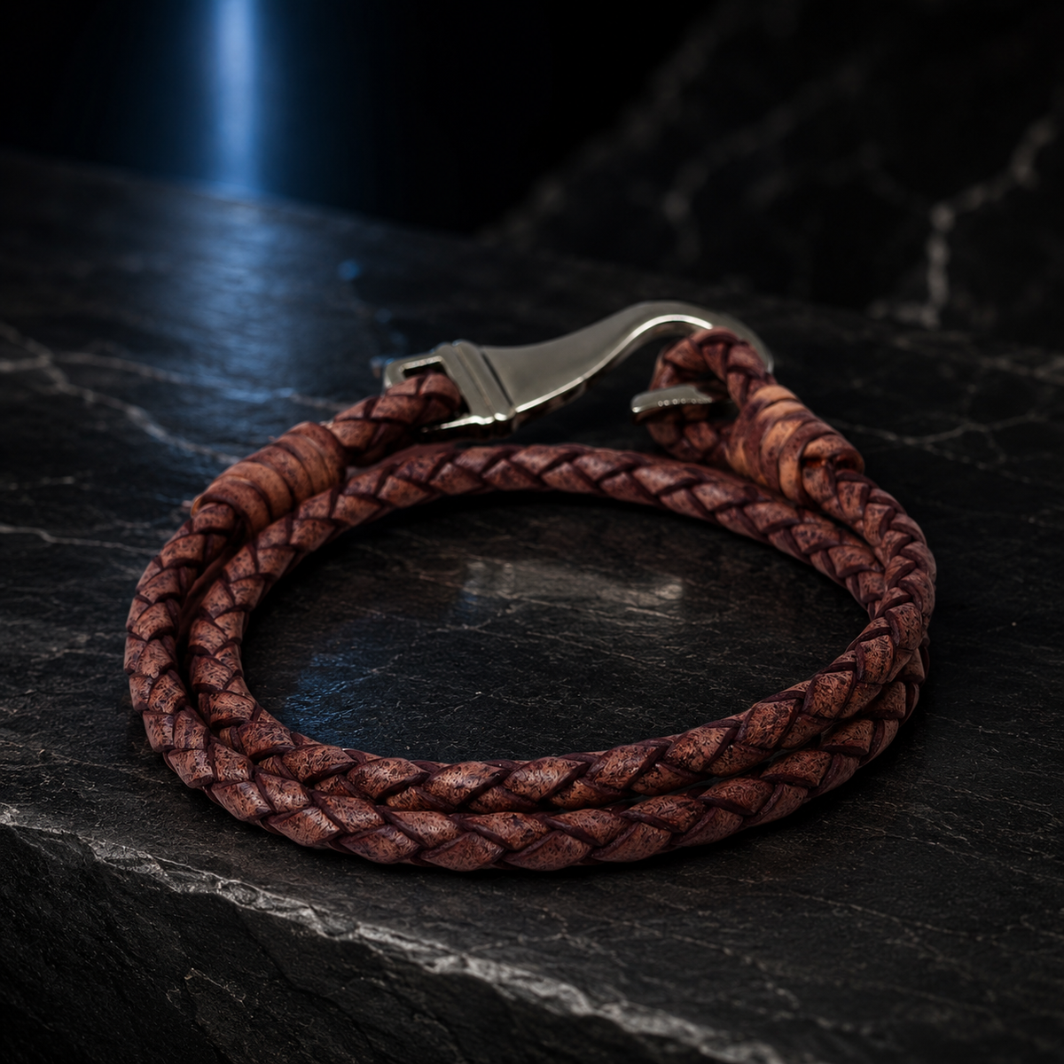 A braided brown leather bracelet with a silver-toned metal lobster clasp, displayed on a dark textured slate surface.